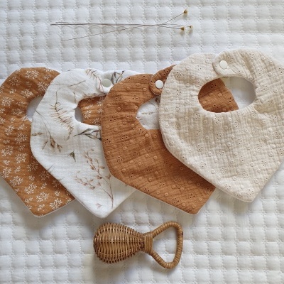 Quatro babetes de tecido coloridos em tons terra e creme com texturas e padrões florais, sobre superfície branca acolchoada, com chocalho de material natural ao lado