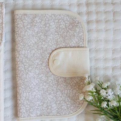Carteira de tecido bege com padrão floral branco e fecho de aba com botão de pressão sobre superfície branca matelassada