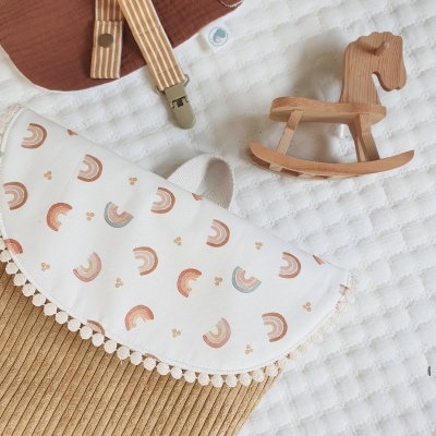 mochila infantil castanha com aba estampada, cavalo de baloiço em madeira, babete castanho e prendedor de chucha às riscas numa superfície branca texturada