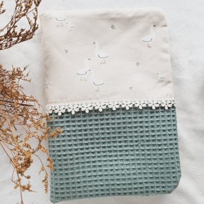 Toalha ou capa de tecido bege com gansos brancos, parte inferior verde textura waffle, detalhe renda branca, sobre fundo branco com ramos secos