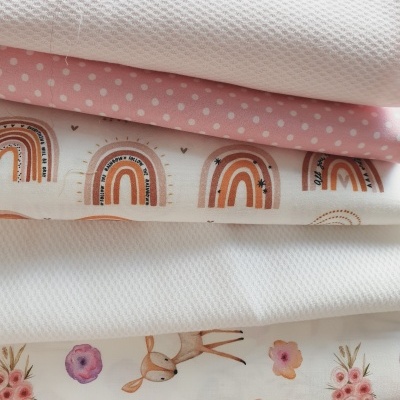 Vários tecidos empilhados com padrões de bolinhas, arco-íris e ilustrações de veado e flores em tons suaves.