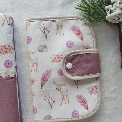 Carteira confeccionada em tecido branco com desenhos de cervos, ouriços e flores em tons rosas e lilás, com aba lilás e botão de pressão, sobre fundo branco e plantas decorativas.