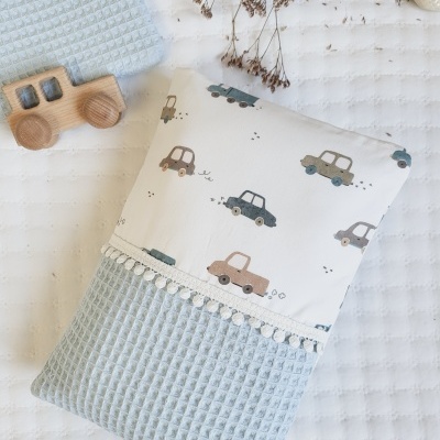 Travesseiro com estampa de carros e tecido azul em padrão colmeia sobre roupa de cama branca com textura, com brinquedo de madeira e flores secas ao lado.