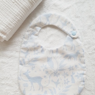 Babete de bebé branco com padrão azul claro de animais e folhas, com fecho de pressão azul, sobre toalha branca.