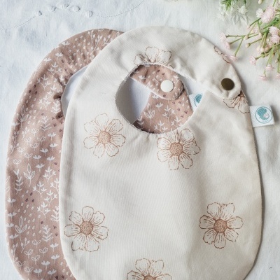 Dois babetes de algodão com padrões florais, um bege com flores castanhas e outro castanho com folhas brancas, sobre pano branco com flores e raminhos floridos ao lado.