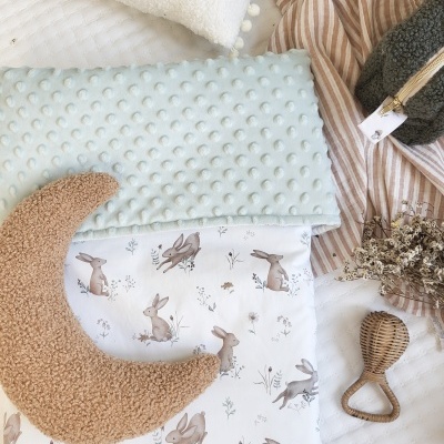 Roupa de cama infantil com padrão de coelhos, manta azul claro, almofada lua castanha e chocalho de madeira.