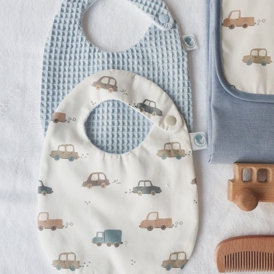 Babetes para bebé azul claro e branco com padrão de carros, cobertor azul e pente de madeira numa superfície branca.
