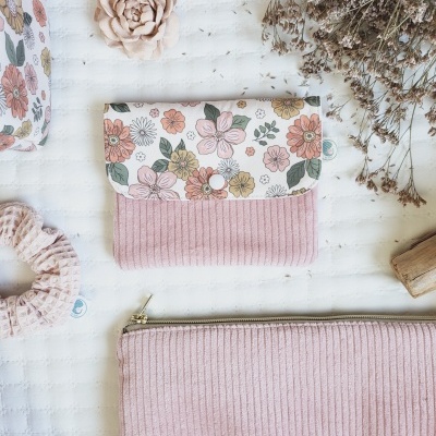 Carteira e bolsa de veludo rosa com padrão floral e botão branco, sobre superfície branca com flores secas