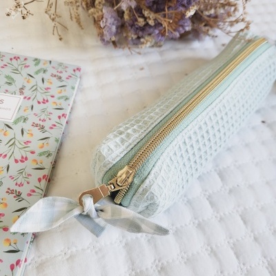 Estojo verde-água matelassê com fecho dourado e fita, bloco de notas floral e flores secas roxas.