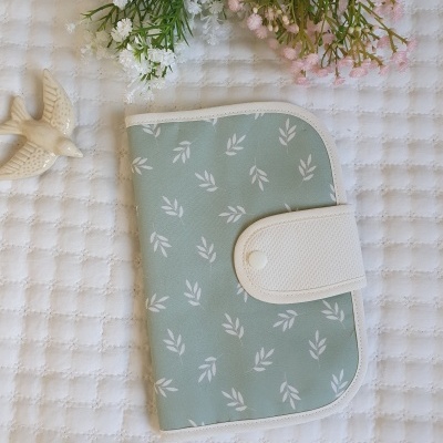 Carteira verde claro com padrão de folhas brancas e fecho branco, sobre tecido branco texturizado com flores e pássaro decorativo