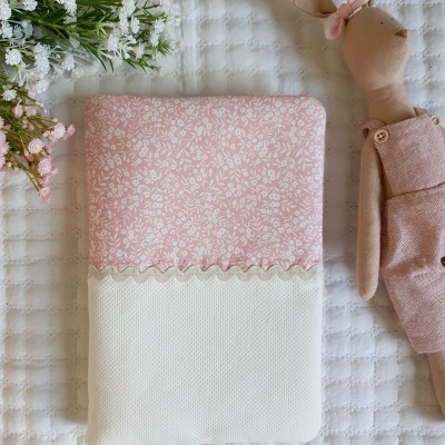 Cobertor rosa e branco com padrão floral, boneca de pano vestida de rosa e flores sobre superfície branca texturada.