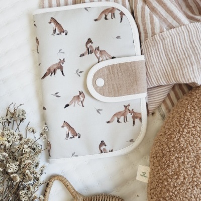 Capa creme com raposas e fecho branco cercada por tecido riscado, ramo seco, chocalho de vime e almofada castanha.