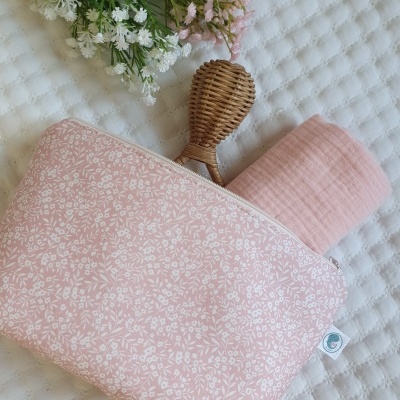 Estojo rosa com padrão branco, pano rosa e objeto trançado sobre superfície branca acolchoada e flores ao fundo