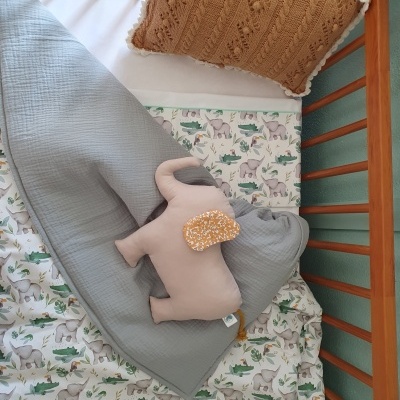 Berço com roupa de cama estampada de elefantes, cobertor cinzento, almofada tricotada castanha e peluche de elefante rosa.