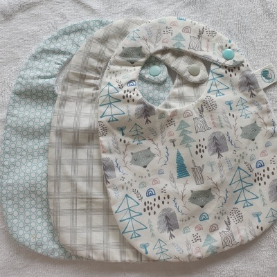 Três babetes de bebé em tecidos com diferentes padrões e fechos de pressão azul.