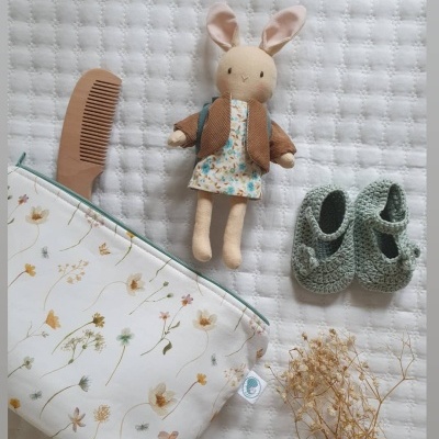 Boneco de coelho de tecido com roupa, pente castanho, sapatinhos verdes de tricô, saco branco com padrão floral e flores secas sobre manta branca