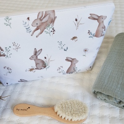 Necessaire branca com padrão de coelhos, escova de cabelo de madeira e tecido verde sobre colchão branco