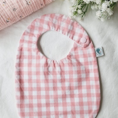 Babete rosa com padrão xadrez e fecho de mola sobre tecido branco com flores e pano rosa com flores