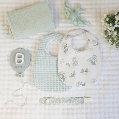 Conjunto azul claro para bebé com babetes, pano, pendente com letra B, presilha e figura de pássaro sobre manta branca