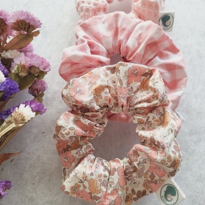 Três scrunchies em tons de rosa com padrões floral, quadriculado e textura rede, com etiqueta branca e logo verde.