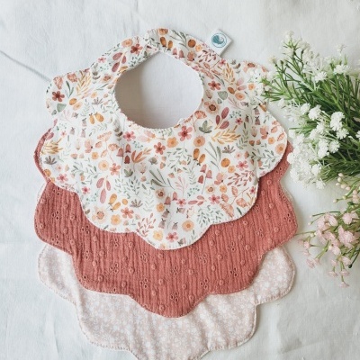 Três babetes em tecido com formato de pétalas, um floral colorido, um rosa texturizado e um rosa claro com padrão floral branco, sobre fundo branco e flores artificiais.