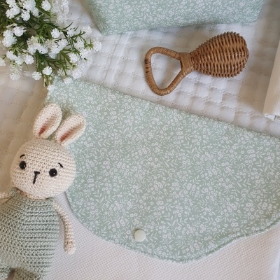 Envelope de tecido branco com aba verde clara e padrão de folhas, brinquedo coelho de crochê, flor branca e chocalho de madeira