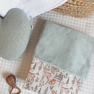 Almofada verde, manta com padrão de veados e cogumelos, mordedor de madeira e cesto de vime sobre tecido branco