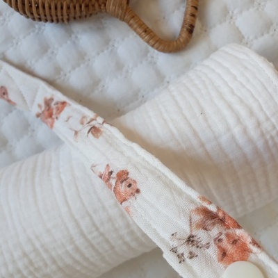 Rolo de tecido branco, faixa floral com botão e objeto de palha trançada sobre tecido acolchoado branco