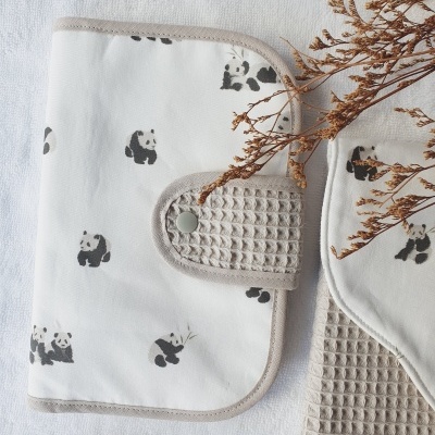 Produto de tecido branco com pandas pretos, aba bege com botão, pente de madeira com gravura 'By Mom' e ramos secos