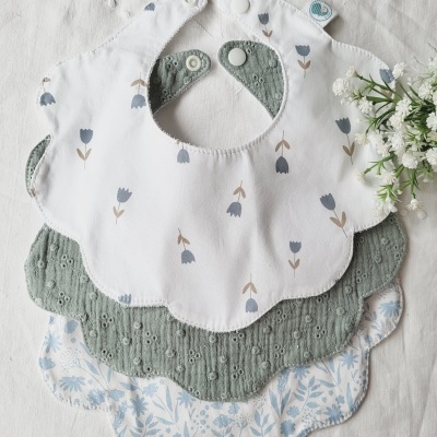 Três babetes infantis com padrões florais e bordas onduladas em branco, verde e azul claro sobre pano branco e flores ao lado.