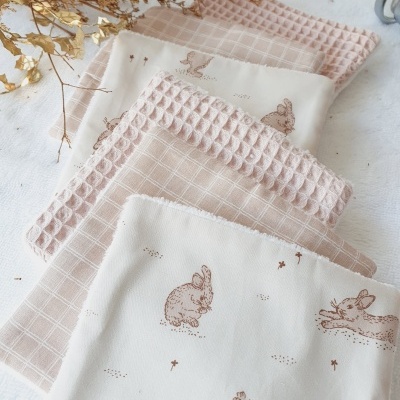 Panhos de cozinha rosa e bege com desenhos de coelhos e texturas variadas, sobre fundo branco com ramos secos.