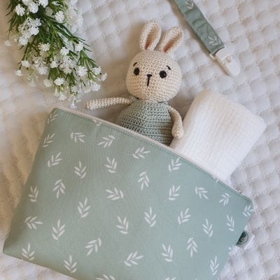 Bolsa verde-água com padrão branco segurando boneco de crochê e toalha branca, sobre fundo acolchoado branco.