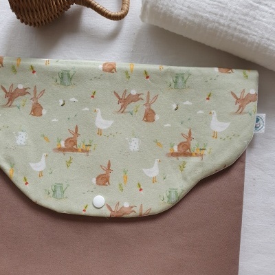 Mala castanha com aba decorada com coelhos e patos, pano branco e maraca em vime.