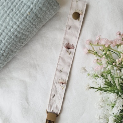 Clip de chupeta com padrão floral em fundo branco ao lado de toalha cinza e flores
