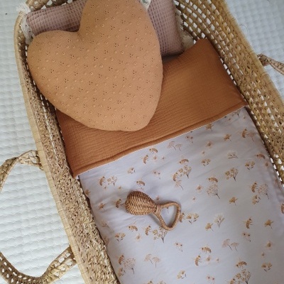 Berço de palha com almofadas castanhas e lençol branco com estampas florais.