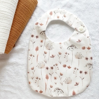 Babete branco com padrão de flores castanhas e rosa colocado sobre tecido branco com duas mantas dobradas ao lado