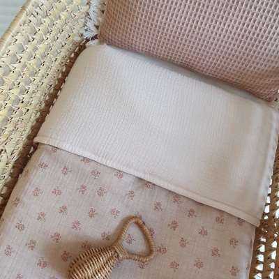 Berço de vime com colchão coberto por lençol branco e cobertor floral, almofada castanha e chocalho de vime