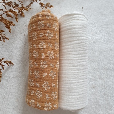 Dois rolos de tecido, um castanho com padrão floral branco e outro branco, sobre superfície clara com ramos secos ao lado.