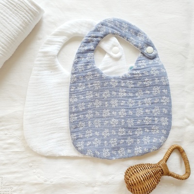 Dois babetes para bebé, branco e azul com flores, e um chocalho em ráfia.