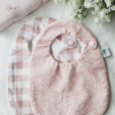 Dois babetes para bebé rosa com padrões e fechos de pressão ao lado de flores brancas