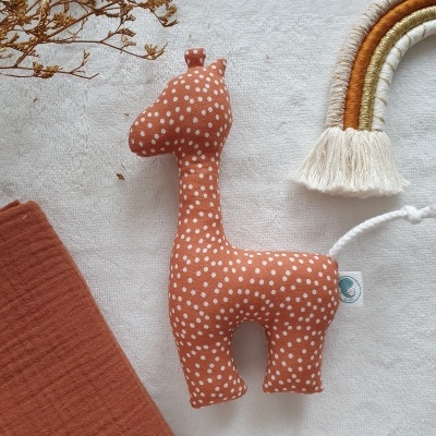 Brinquedo de girafa em tecido castanho com pintas brancas sobre tecido branco, com pano castanho e decoração de arco-íris trançado ao lado.
