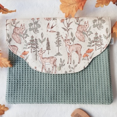 Bolsa de tecido azul esverdeado com aba estampada com animais e plantas, sobre fundo branco com folhas secas e pente de madeira