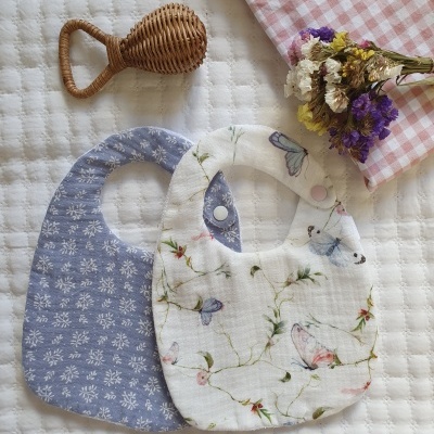 Dois babetes de bebé em tecido com padrões florais e de borboletas sobre tecido acolchoado branco, com chocalho de vime e ramo de flores secas ao lado