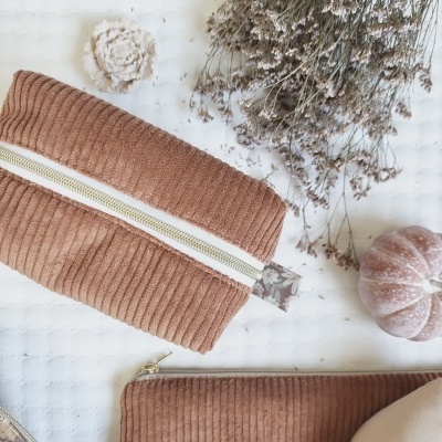 Bolsa e estojo castanhos em tecido canelado com fechos de correr sobre superfície branca com flores e abóbora.