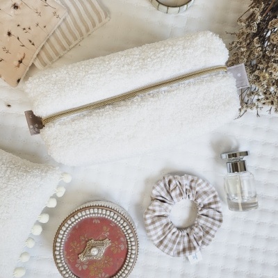 Estojo branco felpudo com fecho dourado sobre tecido branco com padrões, scrunchie xadrez, frasco de vidro, rolo metálico, saco de pano com flores, almofada branca com pompons e prato decorativo vermelho e dourado.
