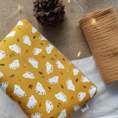 Tecido amarelo com padrão de ouriços brancos, manta castanha, pinha e luzes decorativas sobre fundo de tecido.