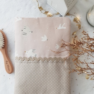 Capa de tecido bege e rosa com padrões de aves e pontos, decorada com fita ondulada bege, sobre superfície branca com elementos secos