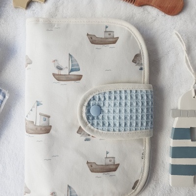 Carteira branca estampada com barcos e gaivotas, fecho azul e pente de madeira 'By Mom' sobre tecido branco