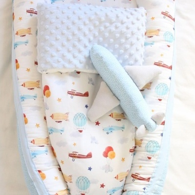 Ninho de bebé com padrão de balões e aviões, almofada cinza texturizada e brinquedo avião azul