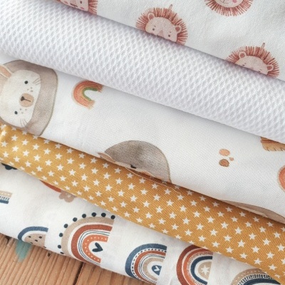 Conjunto de tecidos infantis com padrões de leões, estrelas, arco-íris e animais em várias cores sobre mesa de madeira.